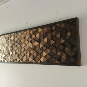 May include: A wooden wall art piece with a geometric pattern of triangles in shades of brown, beige, and white. The triangles are arranged in a gradient pattern, with the lightest shades at the top and the darkest shades at the bottom. The piece is framed in a dark brown frame.