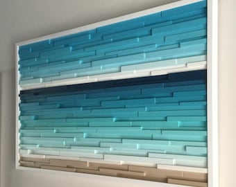 May include: A wall art piece made of wood with a gradient of blue, teal, and white colors, resembling an ocean scene. The wood is arranged in a horizontal pattern, creating a layered effect. The piece is framed in white.