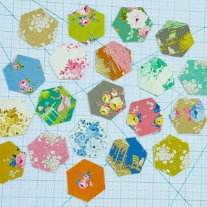 May include: A collection of 17 hexagon-shaped fabric scraps in various colors and patterns, including floral, geometric, and abstract designs. The fabric scraps are arranged on a white gridded surface.