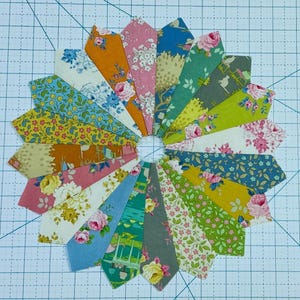 Könnte beinhalten: Ein farbenfroher Stoff-Patchwork-Block mit einem kreisförmigen Design. Der Block besteht aus 18 einzelnen Stoffstücken, jedes mit einem anderen Blumenmuster. Die Stoffstücke sind in einem radialen Muster angeordnet, wobei sich die Spitzen der Stücke in der Mitte des Blocks treffen.