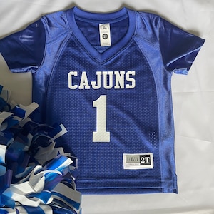 May include: Blue and white football jersey with the word "CAJUNS" and the number "1" on the front. The jersey is a size 2T and is made by B.W. Athletic Wear.