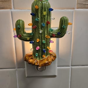 May include: A green ceramic cactus night light with colorful lights. The cactus is plugged into an outlet and is lit up.