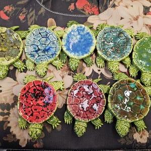 May include: A collection of nine ceramic turtle figurines. Each turtle has a green body and legs, with shells in various colors including blue, green, red, and brown. The shells have textured patterns.