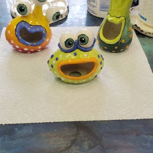 May include: Three ceramic figurines with googly eyes and open mouths. The figurines are painted in bright colors, including green, yellow, orange, blue, and white. The figurines are on a white surface.
