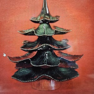 May include: A ceramic treat tree with a layered design, resembling a Christmas tree. The tree is primarily dark green with reddish-brown accents and sits on a round base. The background is a solid red.