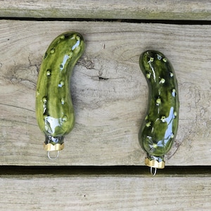 May include: Two green pickle-shaped ornaments with a glossy finish and small white bumps. Each ornament has a gold-colored metal cap and a loop for hanging. The ornaments are displayed on a weathered wooden surface.
