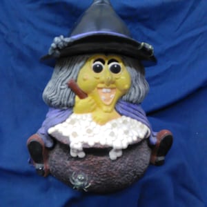 May include: A witch figurine with a black hat, purple robe, and a cauldron filled with white foam. The witch has a yellow face, gray hair, and a pipe in her mouth. A spider is on the side of the cauldron.