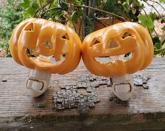 May include: Two orange ceramic pumpkin night lights with a white cord and plug. The pumpkins have a carved jack-o'-lantern face.