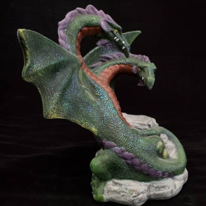 May include: A green two-headed dragon figurine with iridescent glitter, perched on a white rock base. The dragon has purple accents and a curled tail.