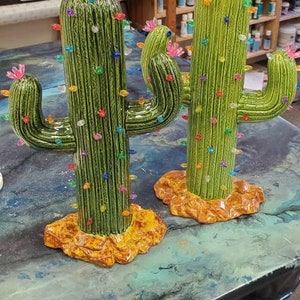 May include: Two ceramic cactus sculptures decorated with colorful lights, resembling Christmas trees. The cacti are green with brown bases and are displayed on a blue and green table.