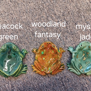 May include: Three ceramic frog-shaped dishes arranged in a row. The left dish is labeled peacock green, the center dish is labeled woodland fantasy, and the right dish is labeled mystic jade. Each frog has a glossy finish and distinct color.