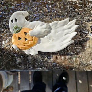 May include: A white ceramic ghost figurine holding a small orange pumpkin with a carved jack-o'-lantern face. The ghost has two black eyes and a small mouth.