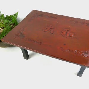 May include: A rectangular low table with a dark reddish-brown lacquered top featuring engraved floral corner patterns and central Japanese characters. The table is supported by four simple black legs, providing a stable base for this piece of furniture.