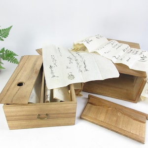 May include: A wooden box with a hinged lid, containing several rolled scrolls of paper with Japanese calligraphy. The box is made of light brown wood and has a brass handle. The scrolls are visible through the open lid.