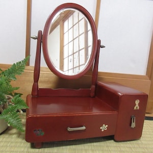 Puede incluir: Un tocador de madera rojo con un espejo ovalado, un cajón y detalles decorativos. El tocador tiene un pequeño compartimento lateral con un diseño floral.