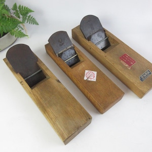 Op de afbeelding: Drie houten handschaven met donkere metalen messen. De schaven zijn in verschillende bruintinten en hebben labels op hun oppervlak. Op de achtergrond is een kleine potplant te zien.