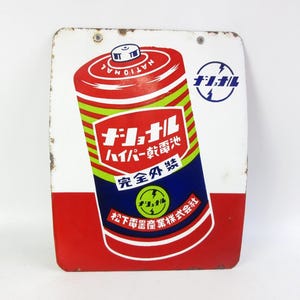 Könnte beinhalten: Ein rechteckiges Metallschild mit einer Grafik einer alten National-Batterie. Das Design umfasst rote, grüne und blaue Farben mit japanischem Text und einem kreisförmigen Logo. Das Schild hat einen weißen Hintergrund mit rotem Rand unten und Rost.