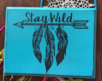May include: A turquoise wooden sign with the text "Stay Wild" in black lettering. The text is centered on the sign and is accompanied by a black arrow with three black feathers hanging from it.