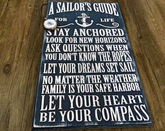 May include: A blue wooden sign with white text that reads "A Sailor's Guide for Life". The sign has nautical-themed sayings, including "Stay Anchored", "Look for New Horizons", "Ask Questions When You Don't Know the Ropes", "Let Your Dreams Set Sail", "No Matter the Weather", "Family is Your Safe Harbor", "Let Your Heart Be Your Compass".