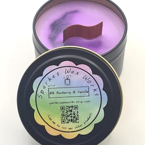 May include: A black tin candle with a purple wax interior and a wooden wick. The lid, placed beside the candle, displays the text "Sparkes Wax Works" and "Blk Rasberry & Vanilla". A QR code and website address are also visible.