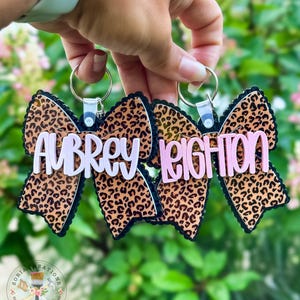 May include: Two bow-shaped keyrings with a leopard print design. The keyrings have black trim and feature the names "Aubrey" and "Leighton" in white and pink lettering. Each keyring has a silver ring attached.