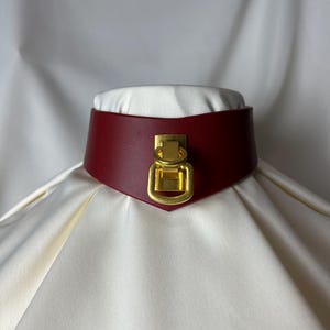 Könnte beinhalten: Ein burgunderfarbener Leder-Choker mit einem goldenen Metallverschluss. Der Choker liegt auf einem weißen Stoffhintergrund.