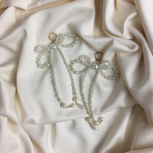 May include: A pair of pearl and gold earrings with a bow design. The earrings are on a white satin background.