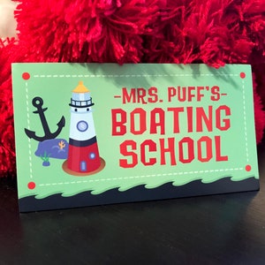 Może przedstawiać: Prostokątny znak z napisem "-MRS. PUFF'S- BOATING SCHOOL" w czerwonych literach. Znak przedstawia kreskówkową latarnię morską, kotwicę oraz zielono-białą kolorystykę. Znak jest umieszczony na czerwonym, rozmytym tle.
