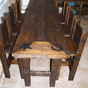 May include: A rustic wooden dining table with a dark brown finish. The table has a rectangular top with a beveled edge and is supported by four sturdy legs. The table top features a carved star and horseshoe design. The table is surrounded by six matching chairs with wooden seats and backs.