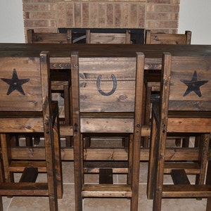 May include: A rustic wooden dining set with a table and six chairs. The chairs have a unique design with a star, horseshoe, or star on the back. The set is made of dark brown wood and has a distressed finish.