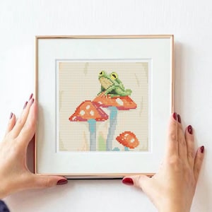 May include: A framed cross-stitch artwork featuring two green frogs sitting on a red and white mushroom. The mushroom has white spots and a green stem.
