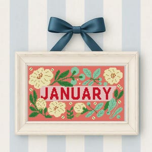 以下が含まれることがあります： 赤い文字で「JANUARY」と書かれた額入りのクロスステッチデザイン。デザインは、クリーム色の花と緑の葉がサンゴ色の背景に囲まれています。青いリボンがフレームの上部に結ばれており、ストライプの壁に掛けられています。