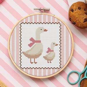 May include: A cross-stitch embroidery hoop showing two geese wearing pink scarves. The geese are beige and brown, set against a white background with a pink and white striped border. The hoop is surrounded by craft supplies and a cup of coffee.