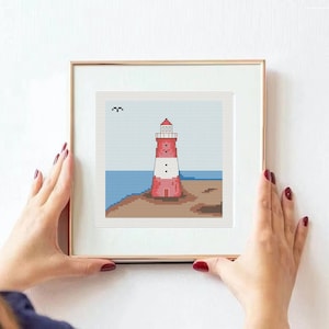 May include: A framed cross-stitch artwork depicting a red and white lighthouse on a sandy shore with blue water in the background.