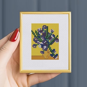 Puede incluir: Una pequeña bordado de punto de cruz enmarcado de un ramo de flores moradas en un jarrón amarillo sobre un fondo amarillo.