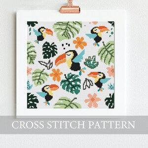 Op de afbeelding: Een ingelijst kruissteekpatroon met een tropisch ontwerp. Het patroon bevat toekans, groene bladeren, oranje bloemen en blauwe accenten op een witte achtergrond. De tekst "CROSS STITCH PATTERN" staat onderaan het frame.
