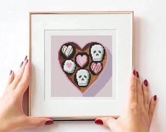 May include: A framed cross-stitch design of a heart-shaped box of chocolates with a skull and crossbones pattern. The chocolates are pink, white, and brown.