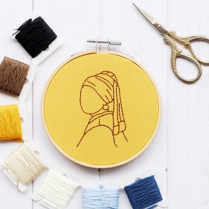 Pode incluir: Um aro de bordado amarelo com um desenho de linha marrom de uma mulher com um brinco de p&eacute;rola. O aro &eacute; cercado por carret&eacute;is de fio de v&aacute;rias cores, incluindo marrom, amarelo, branco, azul e preto.