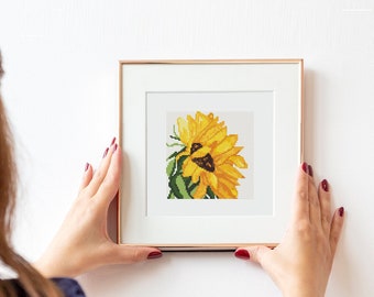 May include: A woman hangs a framed cross-stitch of a sunflower on a white wall. The sunflower is yellow with a brown center and green leaves.