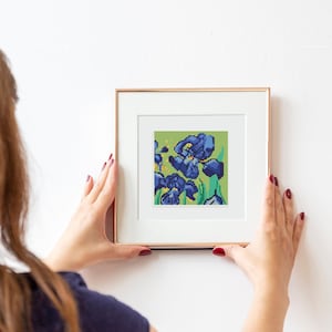 Puede incluir: Una mujer está colgando una impresión enmarcada de una pintura de iris azules en una pared blanca. La impresión está en un marco simple y moderno con un acabado de madera clara.