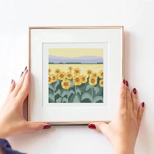 Może przedstawiać: Ramka z haftem krzyżykowym przedstawiająca pole żółtych słoneczników z zielonymi łodygami i liśćmi. Tło przedstawia niebieskie i fioletowe niebo z odległym pasmem gór.