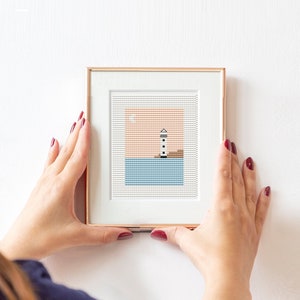 Puede incluir: Una obra de arte de punto de cruz enmarcada que representa un faro blanco con una franja negra sobre un fondo azul y naranja.