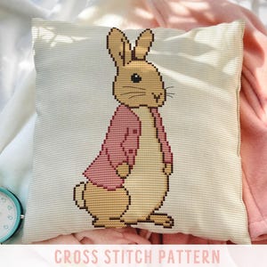 May include: A square, off-white pillow with a cross-stitch pattern of a rabbit wearing a pink jacket. The text "CROSS STITCH PATTERN" is displayed in a pink banner at the bottom of the image. A teal alarm clock is visible in the lower left corner.