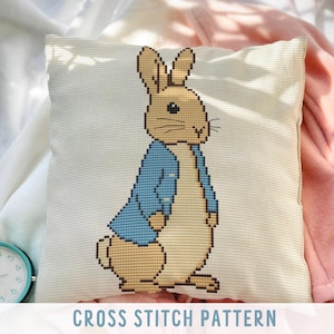 Könnte beinhalten: Ein quadratisches, cremefarbenes Kissen mit einem Kreuzstichmuster eines Hasen mit blauem Jackett. Der Text "CROSS STITCH PATTERN" wird in einem weißen Banner angezeigt. Ein türkisfarbener Wecker und eine rosa Decke befinden sich im Hintergrund.