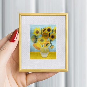 May include: A cross-stitch pattern of a vase of sunflowers in a yellow, blue, and green color scheme. The pattern is framed in a gold frame.