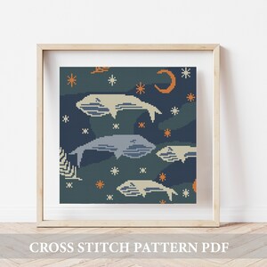 Może przedstawiać: Wzór do haftu krzyżykowego z ciemnoniebieskim tłem, białymi gwiazdami, rogalem księżyca i czterema wielorybami w odcieniach niebieskiego i białego. Wzór jest oprawiony w jasną drewnianą ramę.
