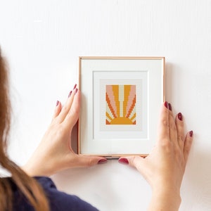 Könnte beinhalten: Ein gerahmtes Kreuzstich-Kunstwerk mit einem Retro-Sonnenstrahlen-Design in Orange-, Pfirsich- und Gelbtönen. Das Kunstwerk befindet sich in einem goldfarbenen Rahmen mit weißem Passepartout und ist bereit zum Aufhängen. Die Person hält das Kunstwerk.
