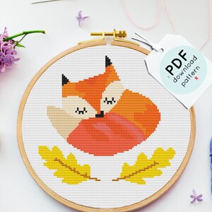 May include: A cross-stitch pattern of a cute orange fox with closed eyes, surrounded by yellow leaves. The pattern is displayed in a wooden embroidery hoop. The image also includes embroidery floss in various colors, a small tag that says "PDF download pattern", and a few small buttons.