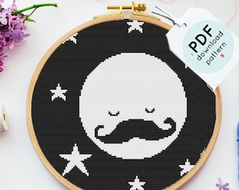 Op de afbeelding: Een zwart-witte kruissteek borduurring met een maan met een snor en sterren. De ring ligt op een wit oppervlak met een label waarop "PDF download patroon" staat en vier klosjes borduurgaren in verschillende kleuren.