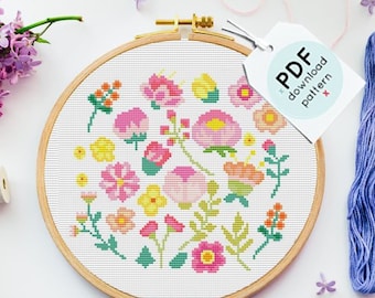 May include: A white embroidery hoop with a colorful floral cross stitch pattern. The hoop is surrounded by four skeins of embroidery floss in shades of blue, pink, and gray. A tag with the text "PDF download pattern" is attached to the hoop.